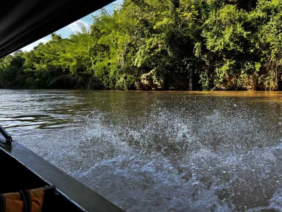 To River Kwai Jungle Rafts by longtail boat. Jungle and river with water splashing next to a boat