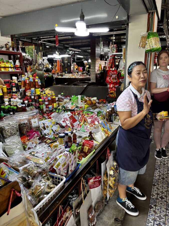 Thai woman standing in front of a colourful market stall, explaining what to buy for a cooking class, with a young western woman looking on