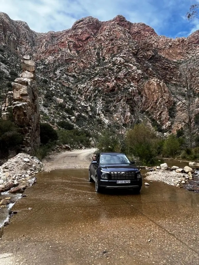 Black car driving through a river, with red rocks and blue sky in the background. Seen when driving back from the Swartberg Pass