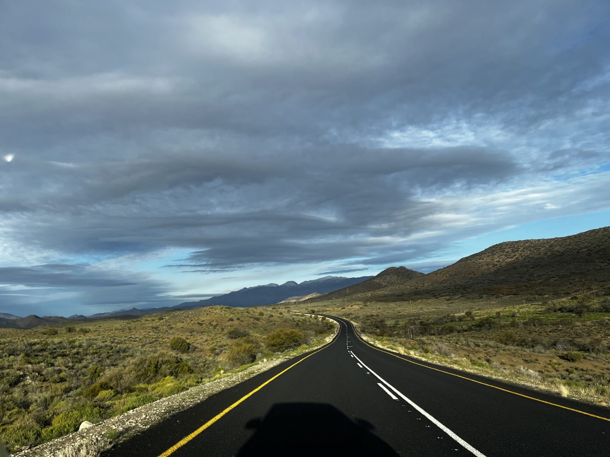 Empty road on the north side of the Swartberg Mountains, between Prince Albert and the Meiringspoort Pass. Black tar road, yellow striped, mountains in the distance, dark clouds.