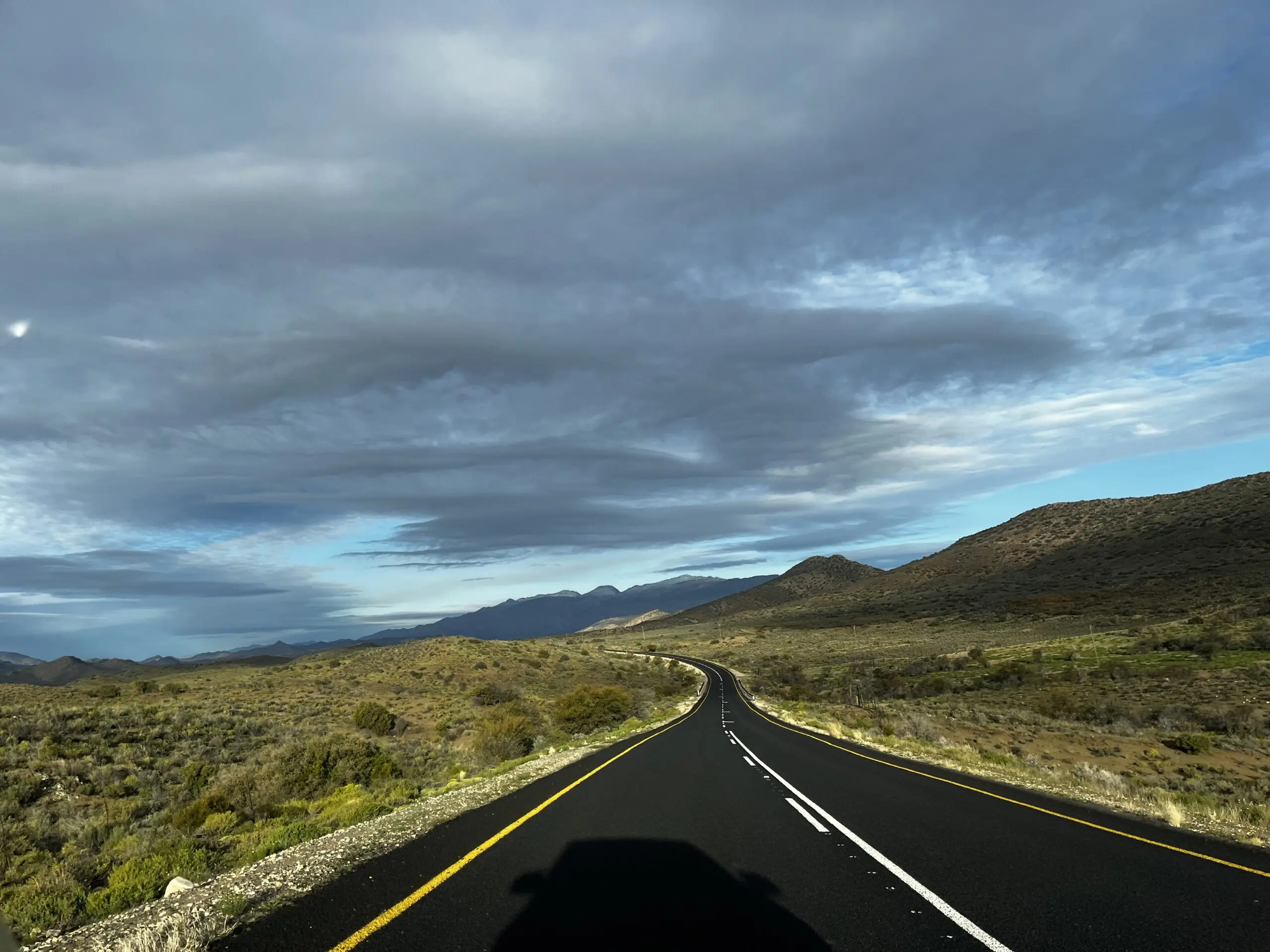 Empty road on the north side of the Swartberg Mountains, between Prince Albert and the Meiringspoort Pass. Black tar road, yellow striped, mountains in the distance, dark clouds.