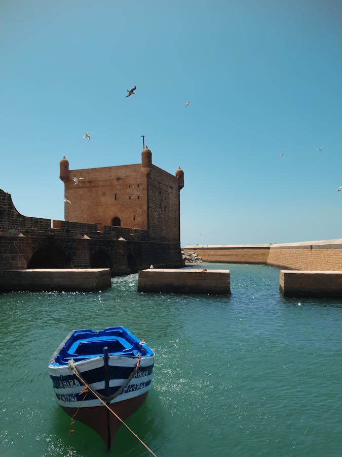 A vibrant scene of a fortress and a blue boat under a clear sky in Essaouira, Morocco. You can visit this on a tour from Marrakech