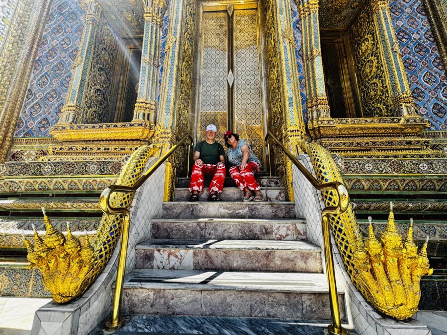 Older couple sitting on the steps of a temple in the Grand Palace in Bangkok. Lots of gold and bling.