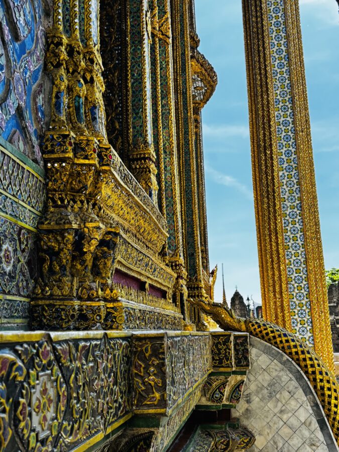 Beautiful detailed side of Thai temple with carvings, tiles and gold decoration in bright colours and bling. Close-up against a bright blue sky.