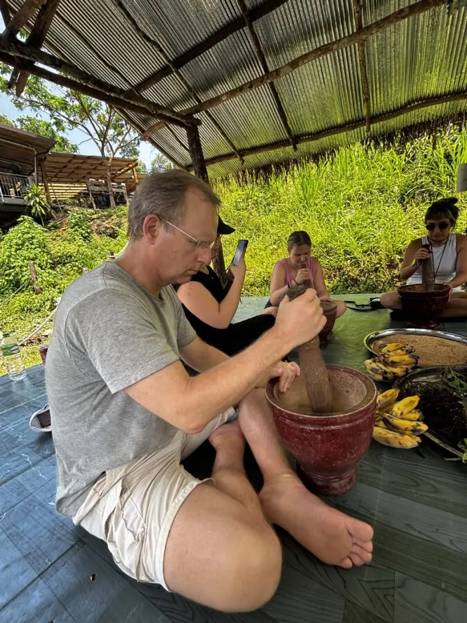 Man in grey t-shirt sitting on the floor with a mortar and pestle, grinding food for elephants 