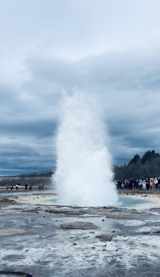 Strokkur geyser erupting with people standing in the background watching and blue water in the foreground. Haukadalur thermal fields, Iceland