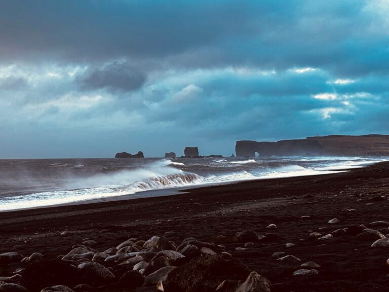 Reynisfjara beach, Iceland, with rocks and black sand in the foreground, crashing waves, stormy skies and basalt formations in the distance.