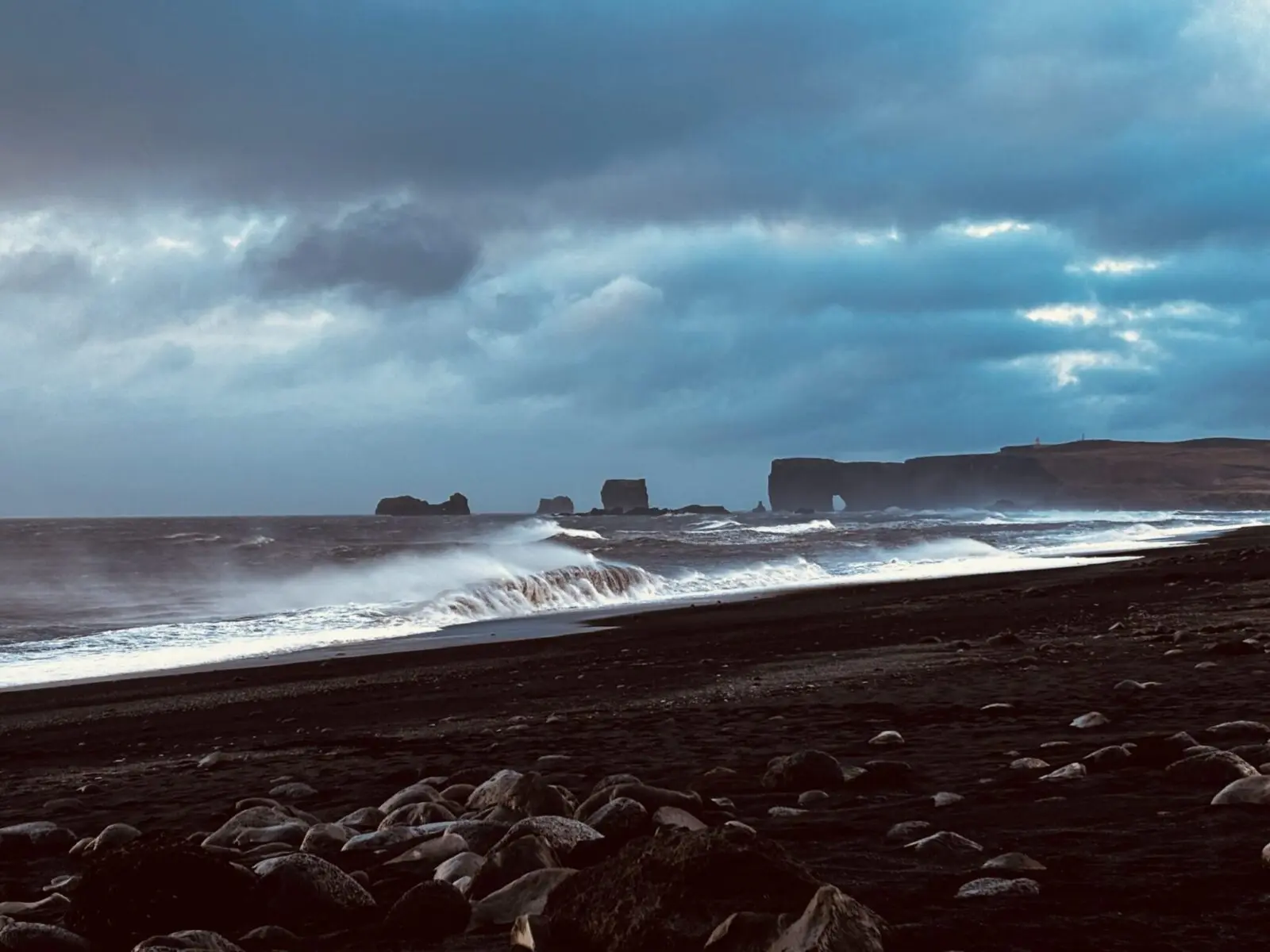 Reynisfjara beach, Iceland, with rocks and black sand in the foreground, crashing waves, stormy skies and basalt formations in the distance.
