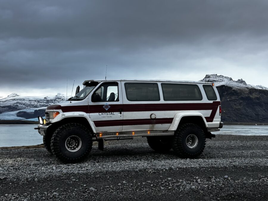 A white minivan with huge tyres standing on a glacier moraine, with lake and mountains in the background. Vattnajökull, Iceland