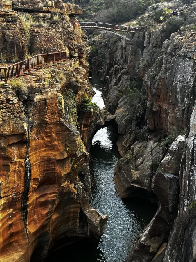 A river seen from above, with red rocks on either side of it, and a metal bridge spanning it. Bourke's Luck Potholes