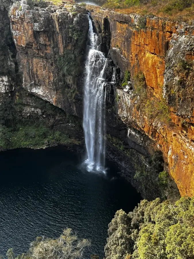 Waterfall dropping straight into a green lake, over red and black rocks. Panorama Route