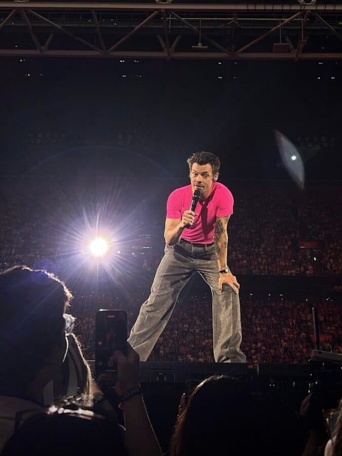Photo: Carmen te Riele. Harry Styles dressed in tan trousers and a pink shirt, listening to a fan in the audience at his concert in Amsterdam. 