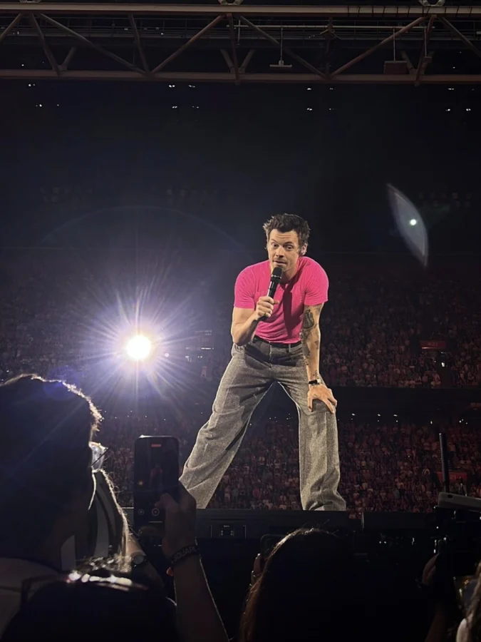 Photo: Carmen te Riele. Harry Styles dressed in tan trousers and a pink shirt, listening to a fan in the audience at his concert in Amsterdam.