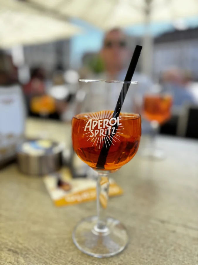 Photo: Tales From The Road Ahead. Shot of an Aperol Spritz cocktail in close-up. In the background, blurred, is another Aperol cocktail and a man in a grey shirt. This is the Aperol travel blog writer Alexandra enjoyed after successfully crossing the border into Germany without a passport. 