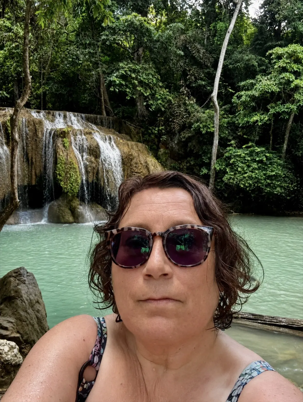 Travel blog writer Alexandra, 57, writing about travel worries, at the first level of Erawan Falls. In a bathing suit, wearing sunglasses and looking slightly dishevellerd.