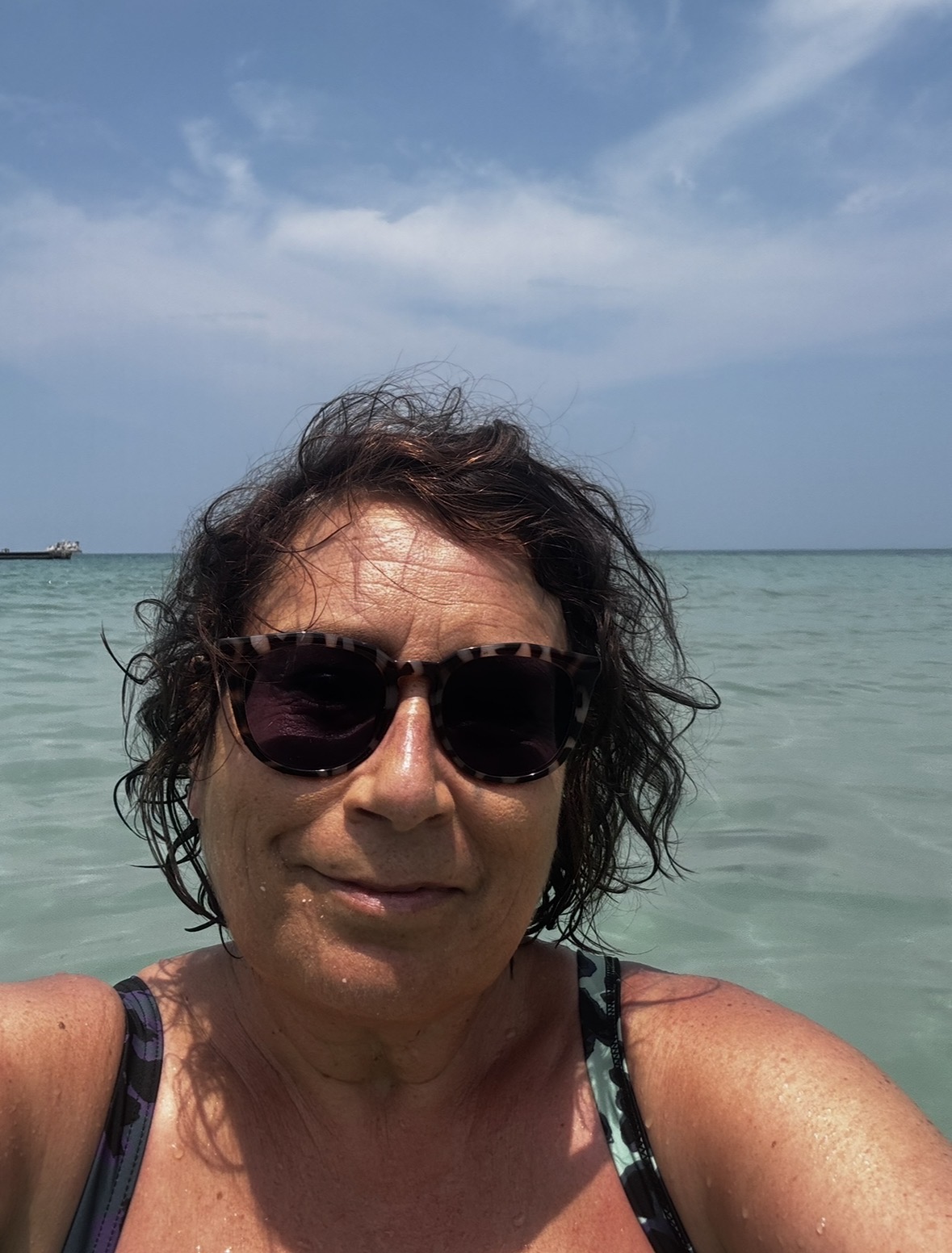 Travel blog writer Alexandra, 57, on a beach. Very tanned, sweaty hair, wearing a bathing suit and sunglasses.