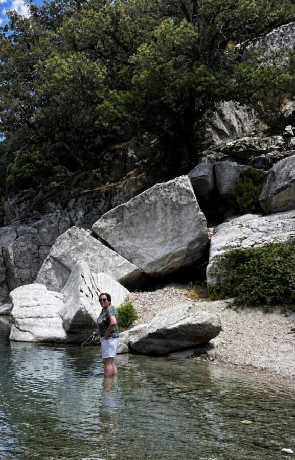 Travel blog writer Alexandra, 57, standing in a clear pool surrounded by big rocks. Back half to the camera, after making it up the mountain despite her worries. 