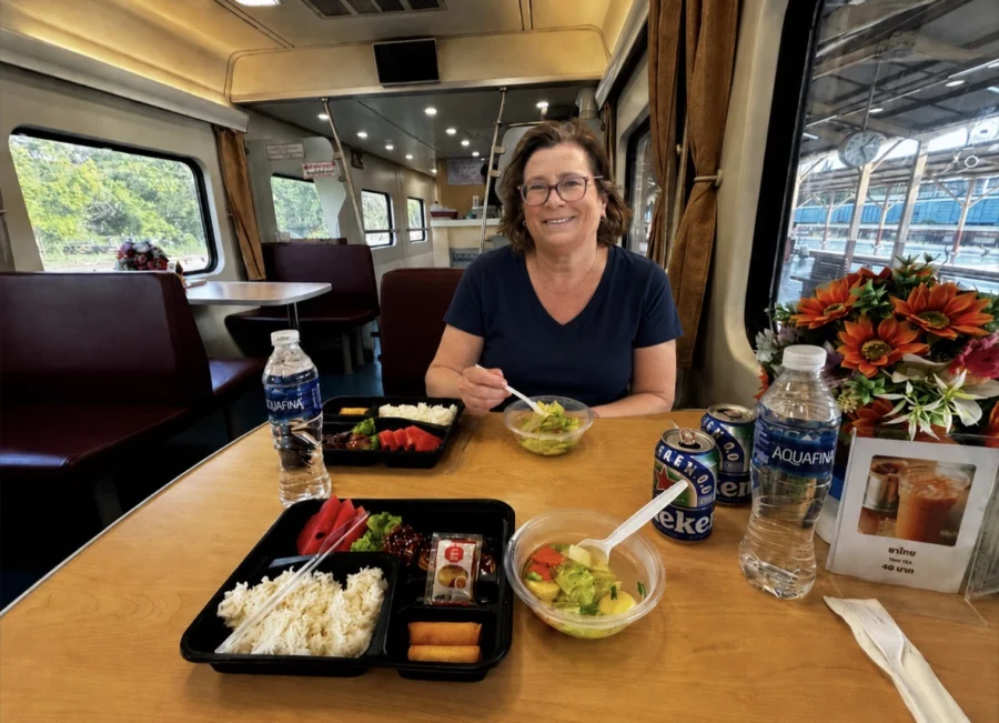 Middle-aged woman sitting at a table in a restaurant carriage on the sleeper train from Chiang Mai to Bangkok, Thailand. This is travel blog writer Alexandra from Talesfromtheroadahead. She has a tray of food, a bottle of water and two empty beer cans in front of her.
