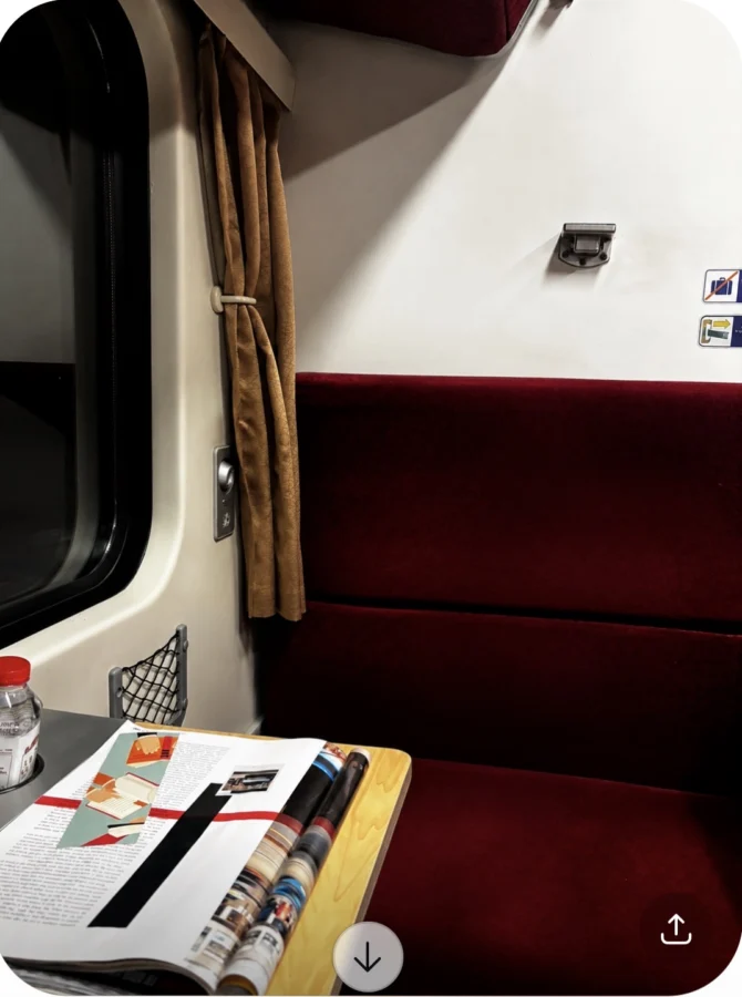A red velvet bench on a train in Thailand. The small table in between holds some magazines and a bottle of water. The curtains are open but it is dark outside.