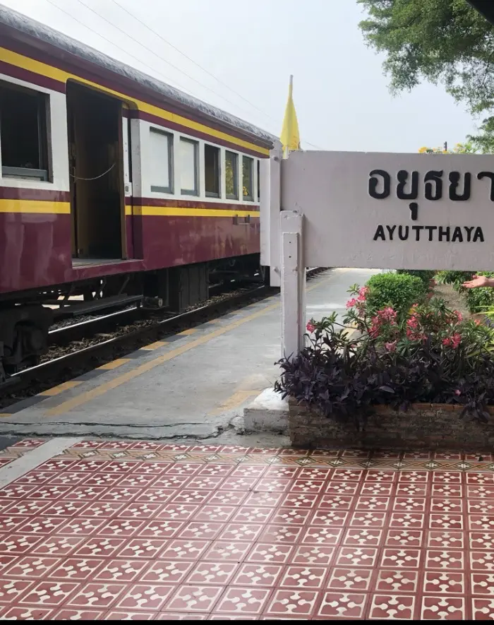 Train in red and yellow standing on a platform. It's the sleeper train from Bangkok to Chiang Mai, Thailand. It has stopped in Ayutthaya; there is a sign with the station name with flowers underneath.