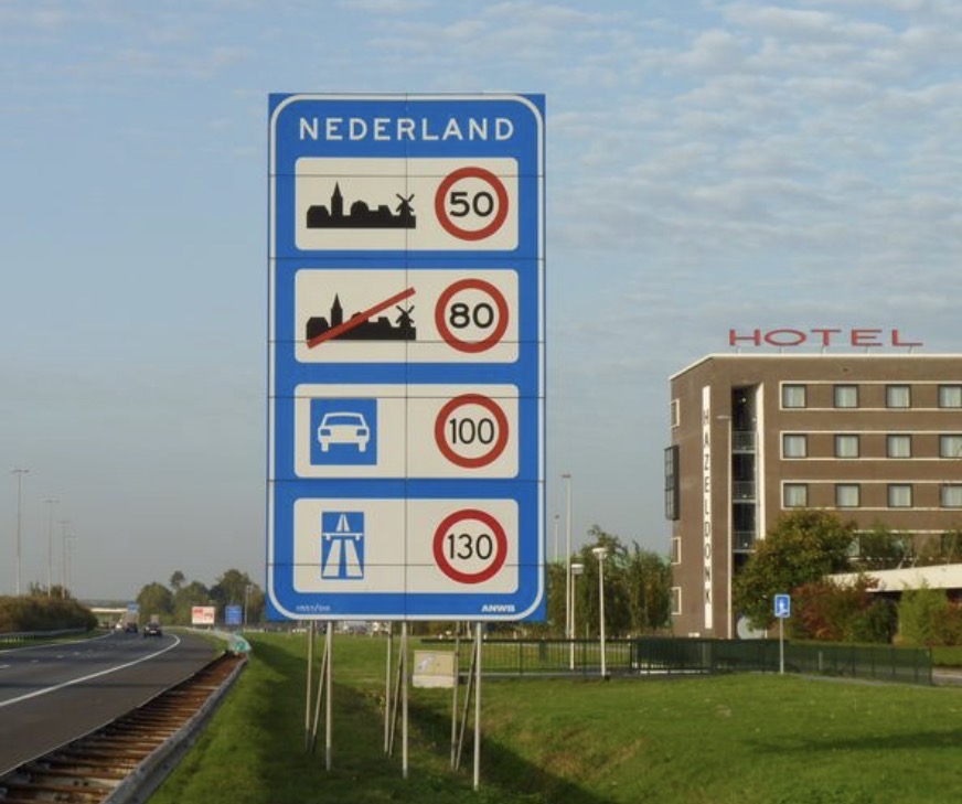 Welcome sign to the Netherlands. Blue sign, maximum speeds indicated, next to a motorway with a hotel on the right of the picture. 